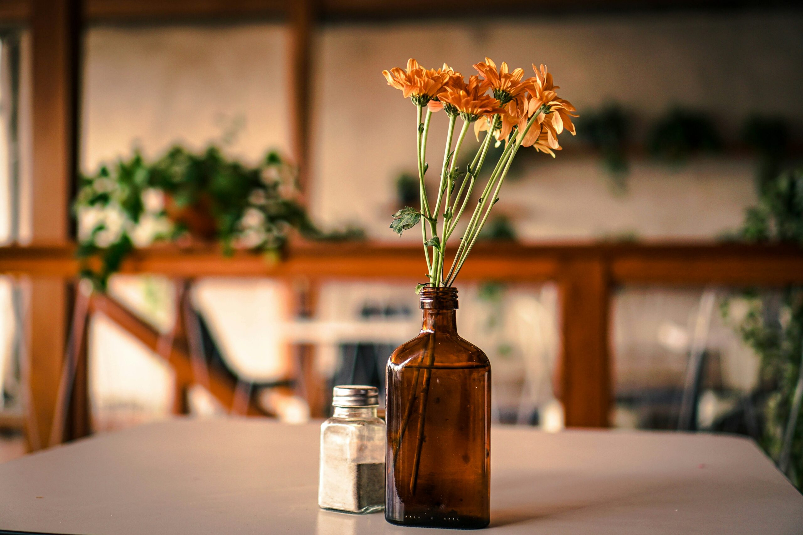Pöytä, jolla on ruskea lasipullo oransseilla kukilla ja pieni maustepurkki, taustalla vihreitä kasveja.